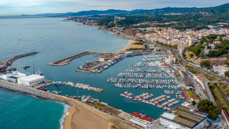 Vista aérea de un puerto deportivo con embarcaciones, rompeolas y zona urbana junto al litoral.