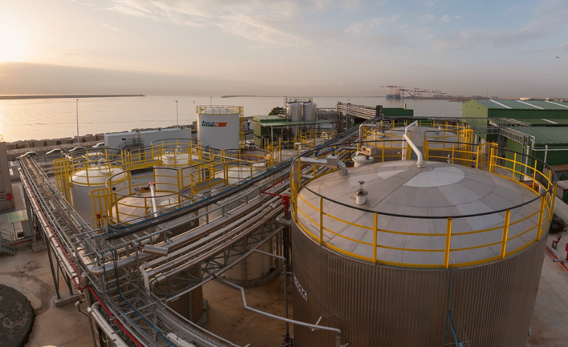 Instalaciones industriales portuarias con tanques de almacenamiento, tuberías y estructuras metálicas junto al mar.
